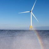 a faraway shot of a wind turbine in the ocean, at the bottom of the image there is mist coming of the water and a rainbow can be seen in the mist